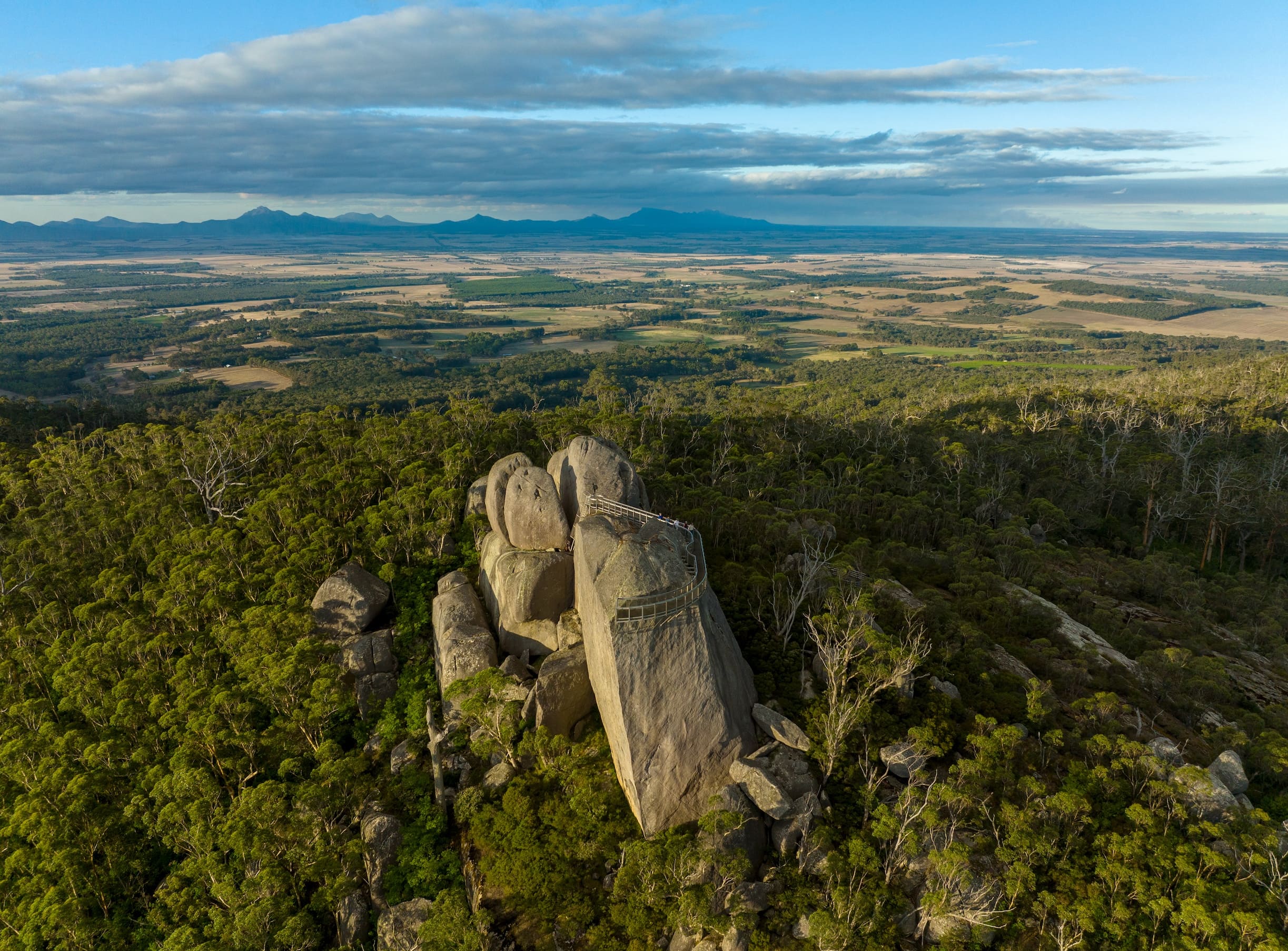 An aerial view of the Granite Skywalk structure built atop massive rock boulders rising from a dense green forest, with a sprawling valley and distant mountains in the background.