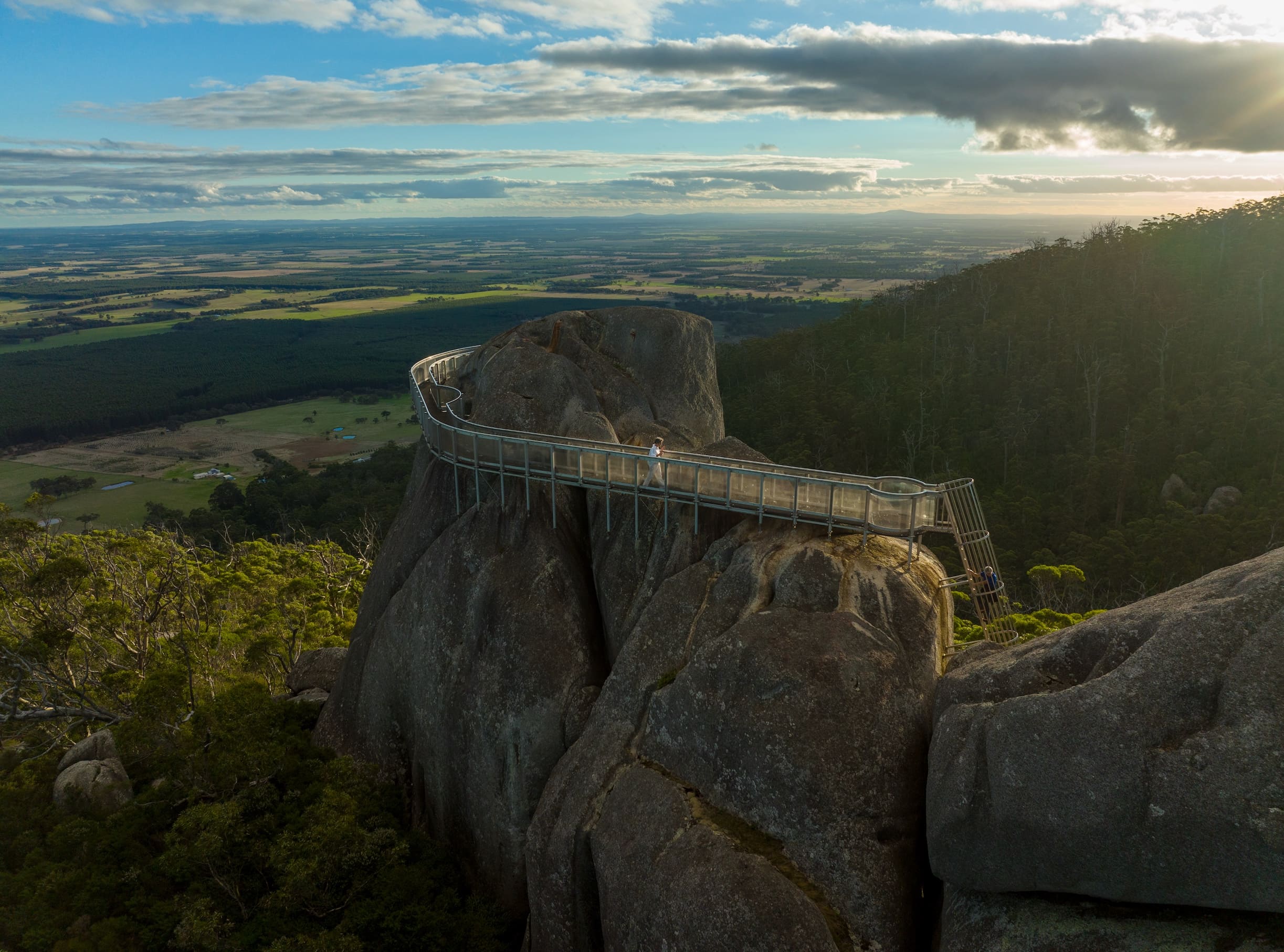 A person walking along the suspended metal and glass walkway of the Granite Skywalk, which wraps around a towering granite peak overlooking a vast agricultural landscape.