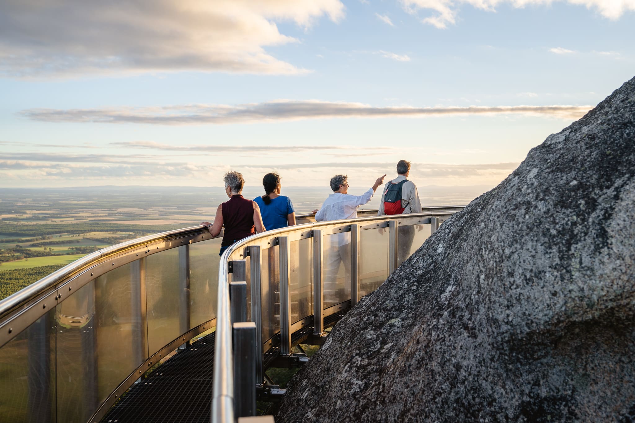 A group of four tour guests standing on the curved observation platform of the Granite Skywalk, looking out over a sweeping panoramic landscape under a partially cloudy sky.