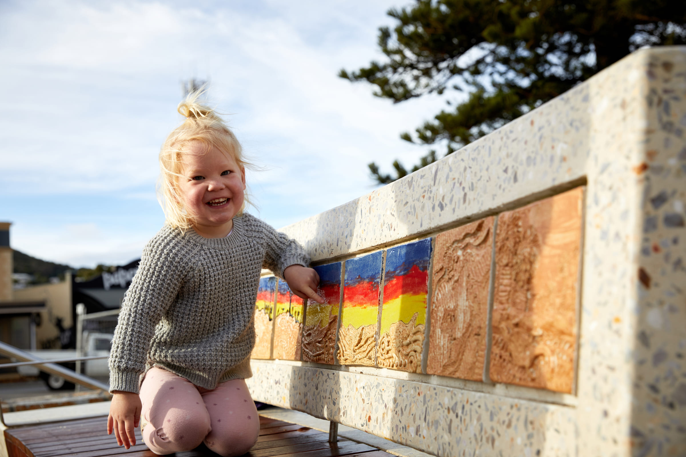 A smiling blonde toddler kneeling on a wooden bench and pointing enthusiastically at brightly colored decorative tiles set into a low concrete wall, interacting with local Albany Arts.